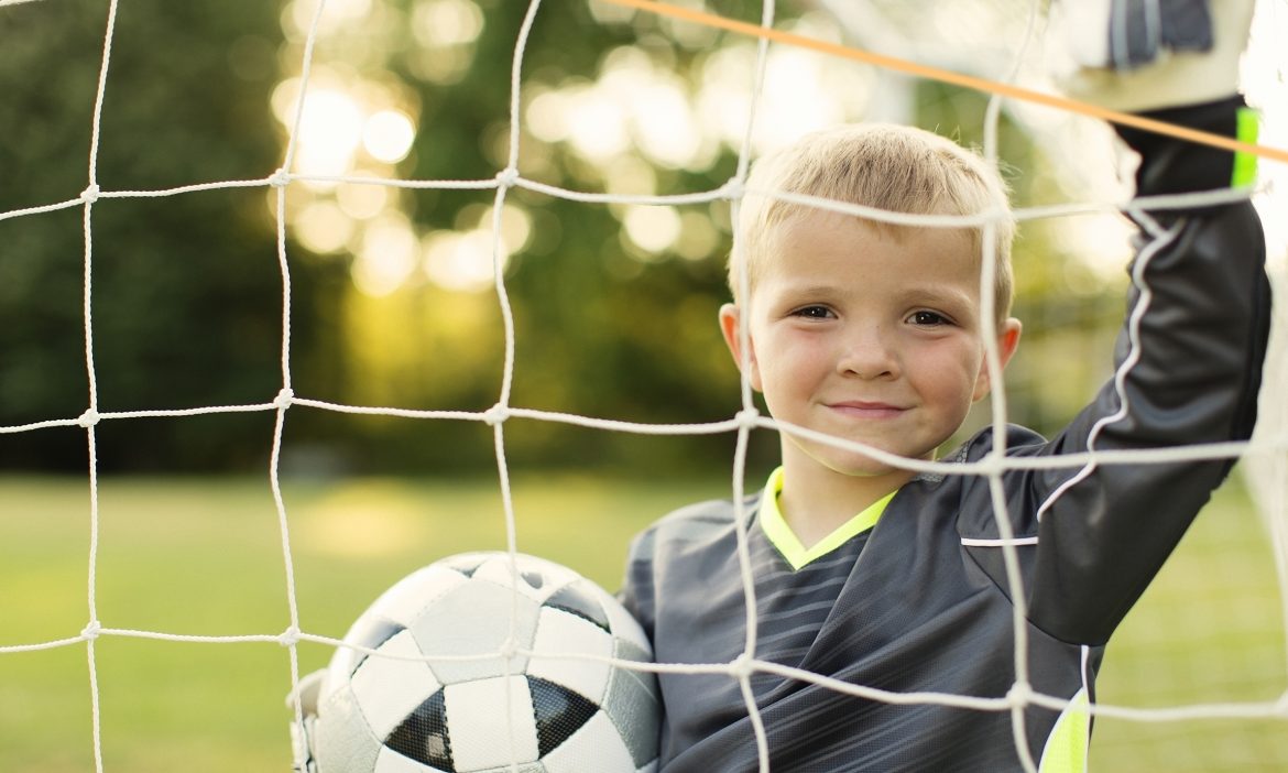 A young boy stands in his soccer uniform holding a soccer ball in the goal while the sun beams through the English trees. He is ready to play little league soccer. Image taken on a summer afternoon.